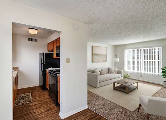 A bright, open living area here at Golden Crest with a cozy carpeted living room featuring a light neutral sofa, wooden coffee table, and large window with vertical blinds, connected to a compact kitchen with warm wood cabinets, dark appliances, and wood-style flooring that creates a clean, welcoming layout.