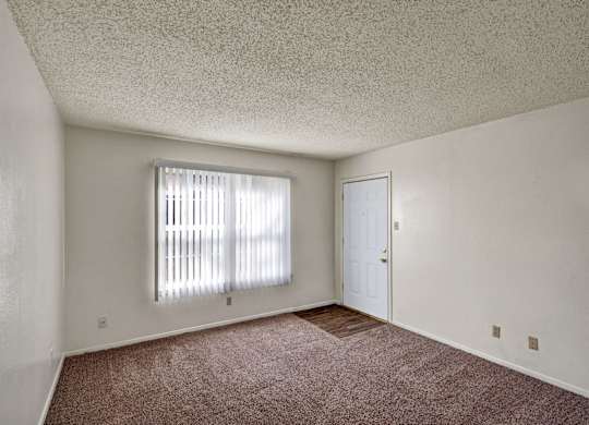 A bright, empty living room here at Golden Crest with plush carpeted flooring, soft neutral walls, and a large window with vertical blinds that lets in natural light, featuring a nearby entry door and a small section of wood-style flooring for a clean, open feel.