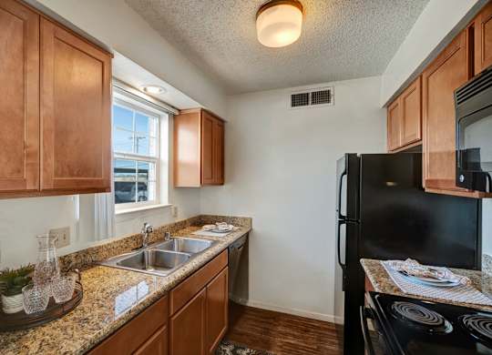 A compact, updated kitchen here at Golden Crest with warm wood cabinets, granite-style countertops, and a double stainless steel sink beneath a window, featuring black appliances and wood-style flooring that create a clean, functional, and inviting space.