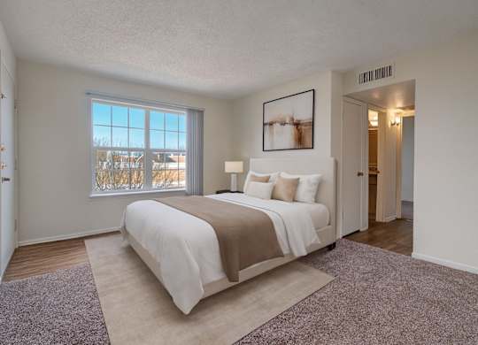 A bright, spacious bedroom here at Golden Crest with plush carpeted flooring, a neatly made bed in soft neutral tones, and a large window with vertical blinds that allows natural light to fill the room, creating a calm and comfortable retreat with easy access to the adjoining hallway and bathroom.