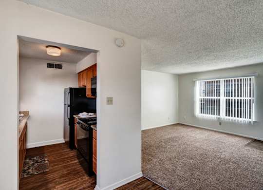 A bright, open living space here at Golden Crest with a carpeted living room and large window with vertical blinds, connected to a compact kitchen featuring warm wood cabinets, black appliances, and wood-style flooring that creates a comfortable and functional layout.
