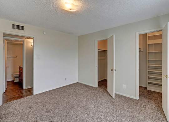 A spacious, empty bedroom here at Golden Crest with plush carpeted flooring, soft neutral walls, and a ceiling light, featuring two closets for ample storage and convenient access to the attached bathroom and hallway.