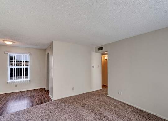 A spacious, empty living area here at Golden Crest with plush carpeted flooring, soft neutral walls, and a large window with vertical blinds, featuring an open layout that connects to a wood-style flooring area and provides easy access to the hallway.