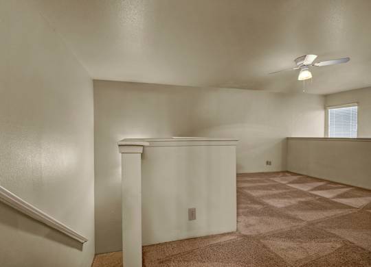 A spacious upstairs loft here at High Plains with soft beige carpet, neutral walls, and a ceiling fan with built-in lighting, featuring an open railing overlook and natural light from a window that creates a flexible, comfortable bonus living space.