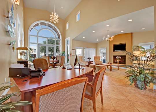 A welcoming community lounge here at High Plains with a shared work table, comfortable seating, tall arched windows, and a cozy fireplace with a mounted TV, enhanced by warm lighting and an open layout that creates a relaxed, social atmosphere.