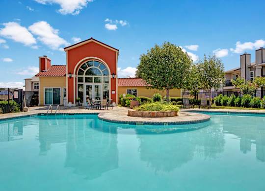 A resort-style swimming pool here at High Plains with clear blue water, surrounding lounge chairs, and a landscaped courtyard, complemented by a clubhouse building and bright skies that create a relaxing, inviting outdoor setting.