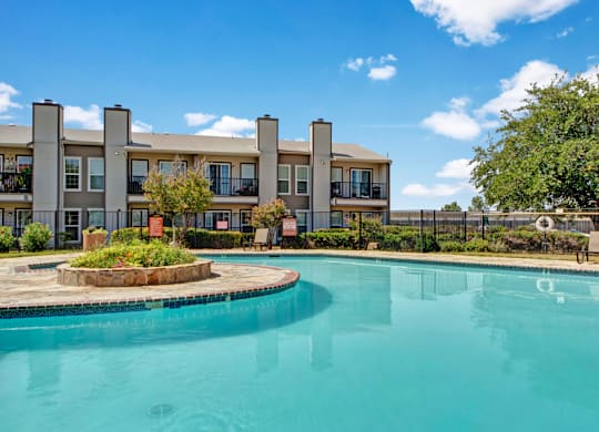 Here at High Plains, a bright outdoor swimming pool with clear blue water sits in a landscaped courtyard, framed by mature trees, fencing, and surrounding apartment buildings with balconies under a sunny blue sky.