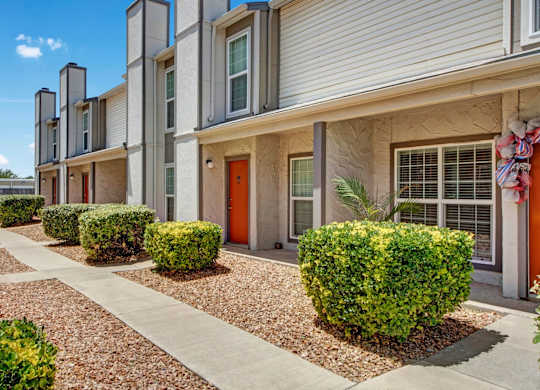 A clean exterior walkway here at High Plains with neatly landscaped shrubs, desert-style gravel beds, and townhouse-style apartment entrances with bright accent doors. The building features large windows and a tidy, welcoming layout under clear blue skies.