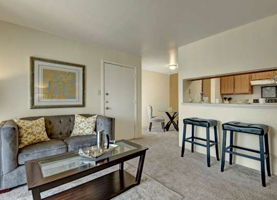 A comfortable living room here at High Plains with soft beige carpet, a cozy sofa, and a glass coffee table, featuring a breakfast bar with seating that opens to the kitchen and a small dining area in the background for a warm, inviting layout.