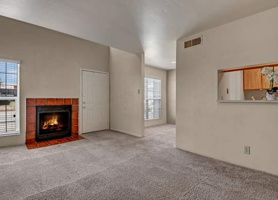 A spacious living room here at High Plains with plush beige carpet, neutral walls, and a cozy fireplace with a tiled surround, complemented by natural light from multiple windows and a pass-through opening to the kitchen for a warm, open feel.
