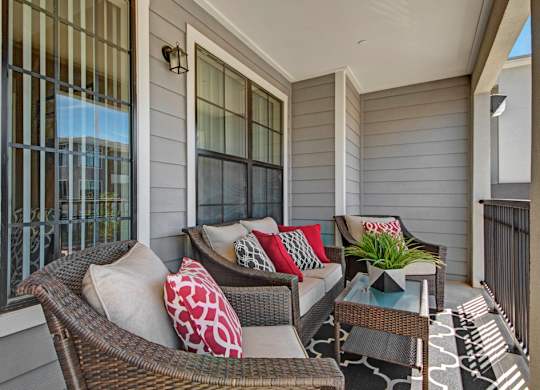 A cozy covered patio here at Le Mirage featuring wicker lounge seating with plush cushions and red accent pillows, a glass-top coffee table with greenery, textured outdoor rug, gray siding, large windows with black trim, and a private railing that creates a comfortable space for relaxing outdoors.