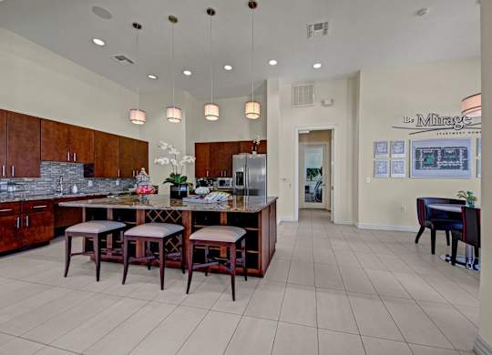 A modern resident lounge kitchen here at Le Mirage featuring a spacious granite island with seating, rich wood cabinetry, stainless appliances, pendant lighting, tiled flooring, and an open layout that creates a welcoming space for gatherings, casual dining, and community events.