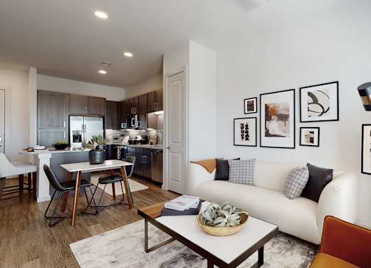 A bright open-concept living room and kitchen here at Magnolia on the Green with wood-style floors, dark cabinets, stainless steel appliances, a small dining table, and a modern sofa with wall art for a warm, stylish feel.