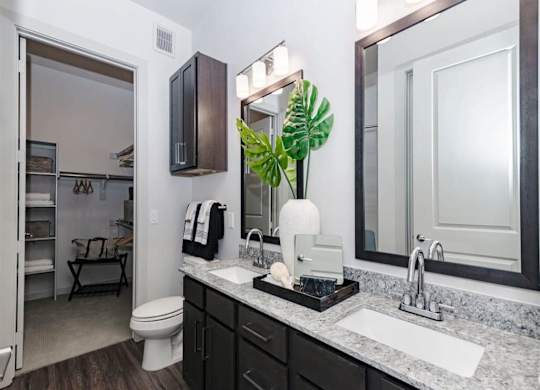 A modern bathroom here at Magnolia on the Green with a double-sink vanity, granite countertop, dark cabinets, framed mirrors with bright lighting, and an open walk-in closet with built-in shelving nearby.