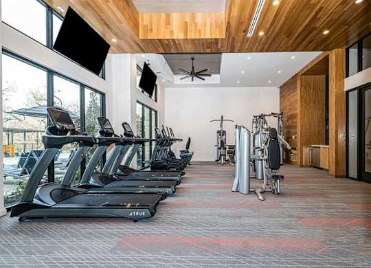 A modern fitness center here at Magnolia on the Green with treadmills, strength machines, high ceilings with wood accents, wall-mounted TVs, and large windows bringing in natural light for an open, energizing workout space.