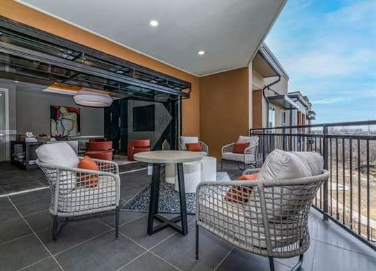A covered outdoor lounge here at Magnolia on the Green with cushioned patio chairs, a round table, tiled flooring, and railing views, connected to an indoor seating area for a relaxed, social atmosphere.