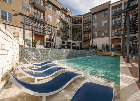 A resort-style pool area here at Magnolia on the Green with in-water lounge chairs, a cascading water feature wall, surrounding apartment buildings with balconies, and sunny deck space for relaxing outdoor enjoyment.