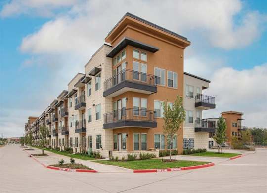 A modern exterior building view here at Magnolia on the Green with multi-story apartments, private balconies, large windows, landscaped sidewalks, and clean architectural lines along a wide corner street.