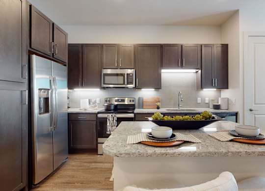 A modern kitchen here at Magnolia on the Green with dark wood cabinets, granite countertops, stainless steel appliances, a center island with seating, and bright under-cabinet lighting for a clean, upscale feel.