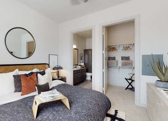 A bright bedroom here at Magnolia on the Green with plush carpet, a large bed with layered pillows, bedside lamps, a round wall mirror, and an open closet and bathroom doorway for a comfortable, connected layout.