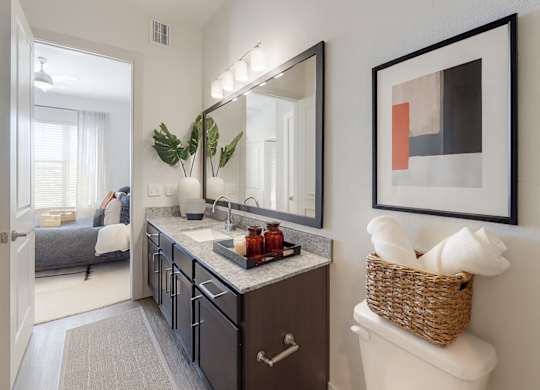 A bright bathroom here at Magnolia on the Green with a dark wood vanity, granite countertop, wide mirror with modern lighting, and stylish decor, connected directly to a nearby bedroom for convenient access.