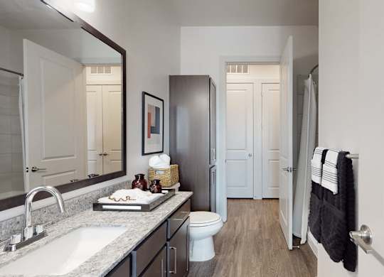 A clean bathroom here at Magnolia on the Green with a dark wood vanity, granite countertop, large mirror, tile shower, and wood-style flooring, creating a bright, modern, and functional space.