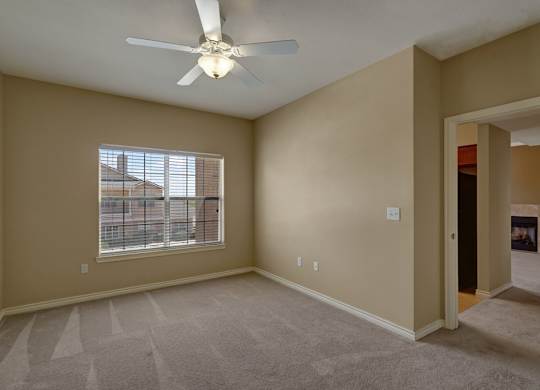 A spacious bedroom here at Mission Green featuring soft neutral walls, plush carpet flooring with clean vacuum lines, a ceiling fan with overhead lighting, and a large window with blinds that brings in natural light, creating a comfortable and versatile space with an open layout that connects easily to the adjacent living area.