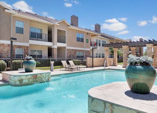 A resort-style outdoor pool area here at Mission Green showcasing clear blue water, a spacious stone pool deck, large decorative planters, shaded pergola columns, lounge chairs, and surrounding apartment buildings under a bright sky, creating a relaxing community space for residents to unwind.