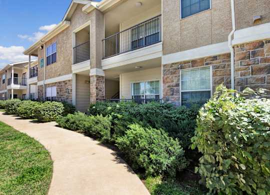 A well-maintained apartment building exterior here at Mission Green showcasing stone and stucco architecture, private balconies, large windows, neatly trimmed landscaping, and a curved concrete walkway bordered by green grass and shrubs beneath a bright blue sky.