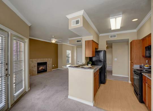 A bright and open living area here at Mission Green showcasing an open-concept layout with a connected kitchen and living space, warm neutral walls, wood-style flooring in the kitchen, soft carpeted living area, a tiled fireplace, ceiling fan, and large windows and glass doors that allow natural light to fill the room.