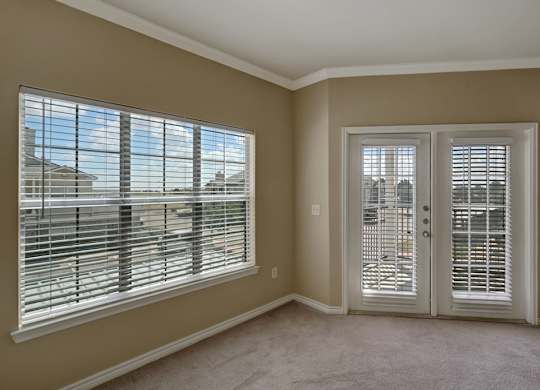 A bright and comfortable living room here at Mission Green showcasing neutral-toned walls, soft carpeted flooring, a wide window with blinds that brings in natural light, and glass patio doors with matching blinds that open to an outdoor area, creating an airy and inviting atmosphere for everyday living.