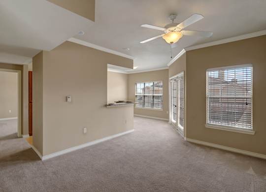 A bright and open living room here at Mission Green featuring plush carpet flooring, neutral wall tones, a ceiling fan with overhead lighting, and multiple windows that bring in natural light, with a pass-through counter to the kitchen and an open layout that connects comfortably to adjacent rooms.