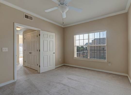 A comfortable bedroom here at Mission Green featuring soft neutral walls, plush carpet flooring, a ceiling fan with overhead lighting, and a large window with blinds that brings in natural light, with double-door closet access and an open doorway that connects conveniently to the rest of the apartment.