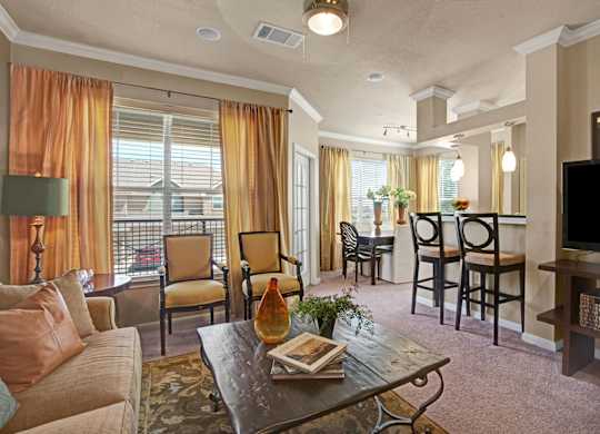 A warm, inviting living room here at Palms at Briarwood featuring plush carpeting, a comfortable sofa with accent pillows, a rustic coffee table, and soft window treatments that frame a private balcony, with an open layout flowing into a dining area and breakfast bar with barstools under modern pendant lighting.