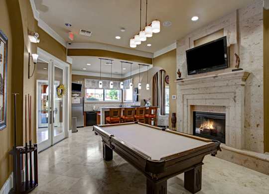 A stylish resident lounge here at Palms at Briarwood featuring a pool table, stone fireplace, mounted television, and bar seating, with pendant lighting, polished floors, and an open layout creating a welcoming space for relaxing, socializing, and entertaining guests.