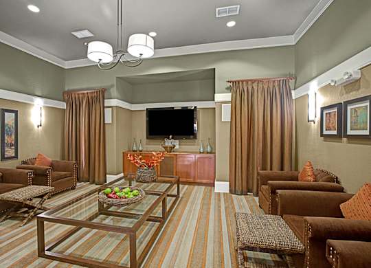 A comfortable resident lounge here at Palms at Briarwood showcasing plush seating, glass coffee tables, a mounted television, and warm neutral tones, with soft lighting, framed wall art, and an inviting layout designed for relaxing, gathering with neighbors, or enjoying casual entertainment.