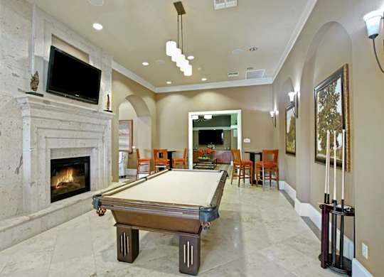 A stylish community game room here at Palms at Briarwood featuring a full-size billiards table, a stone-accent fireplace with mounted television, arched entryways, bar-height seating, pendant lighting, framed artwork, and polished flooring that creates a welcoming space for socializing and recreation.