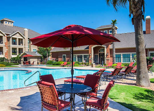 A resort-style outdoor pool area here at Palms at Briarwood featuring a sparkling swimming pool, red cushioned lounge chairs, shaded patio tables with umbrellas, manicured landscaping, palm trees, and surrounding apartment buildings that create a relaxing, inviting setting for residents to unwind.