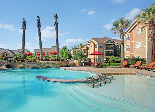 A resort-style pool area here at Palms at Briarwood featuring a sparkling swimming pool with a shallow sun shelf and in-water loungers, tall palm trees, red patio umbrellas, cushioned lounge chairs, stone accents, and surrounding apartment buildings under a bright blue sky.