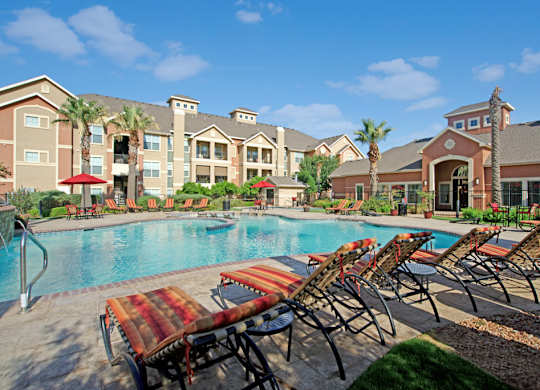 A resort-style pool area here at Palms at Briarwood featuring a large sparkling swimming pool surrounded by cushioned lounge chairs, red patio umbrellas, palm trees, landscaped greenery, and nearby apartment buildings and clubhouse under a bright blue sky.