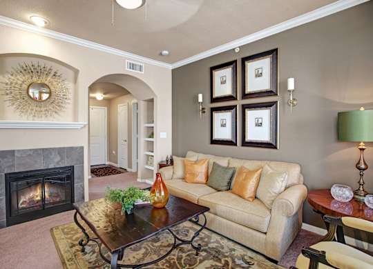 A cozy, well-appointed living room here at Palms at Briarwood featuring plush carpeting, a comfortable sofa with accent pillows, a decorative fireplace with tiled surround, an arched entryway, framed wall art, and a wooden coffee table that creates a warm, inviting space for relaxing or entertaining.