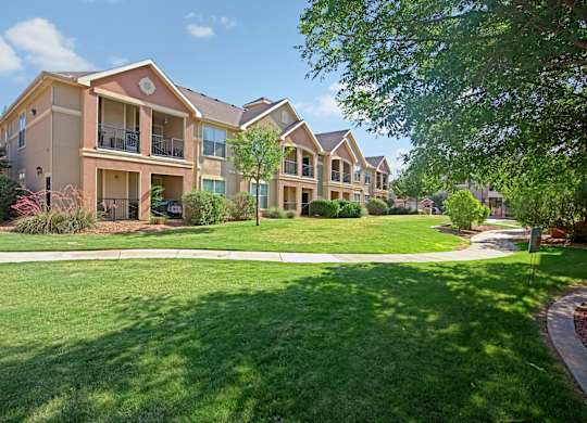 A landscaped apartment exterior here at Palms at Briarwood showcasing multi-story residential buildings with covered balconies, expansive green lawns, mature shade trees, trimmed shrubs, and a curved walking path creating an inviting, park-like community setting under a bright blue sky.