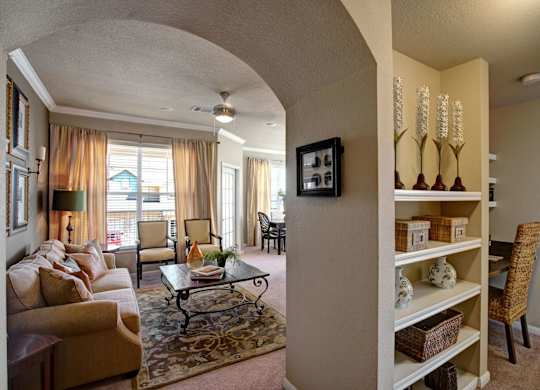 A welcoming, open-concept living room here at Palms at Briarwood featuring plush carpeting, an arched entryway, built-in shelving with decorative accents, a comfortable sofa with neutral pillows, a patterned area rug, and a coffee table, with natural light flowing in from large windows and an adjacent dining area visible beyond.