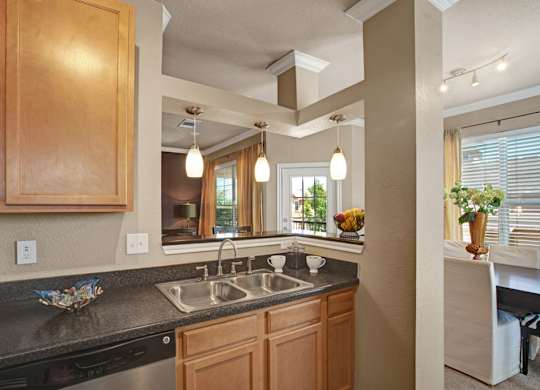 A welcoming kitchen here at Palms at Briarwood featuring warm wood cabinetry, a double-basin sink with a sleek faucet, dark countertops, and pendant lighting above the pass-through, offering an open view into the dining and living areas that creates a bright, connected layout ideal for everyday living and entertaining.