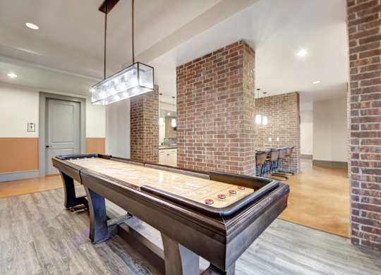 A game lounge here at Sorrel Fairview with a shuffleboard table, brick columns, warm concrete floors, modern rectangular pendant light, and a bar-height counter with stools in the background.