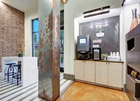 A coffee bar area here at Sorrel Fairview with a self-serve coffee machine, light wood cabinets, dark tile backsplash, brick accent wall, tall metal column, and a white high-top table with black stools.