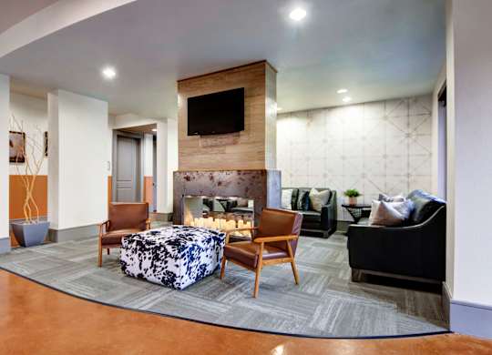 A lounge seating area here at Sorrel Fairview with a modern fireplace wall and TV, brown leather chairs, black sofa, patterned ottoman, and soft neutral finishes, warm and inviting.