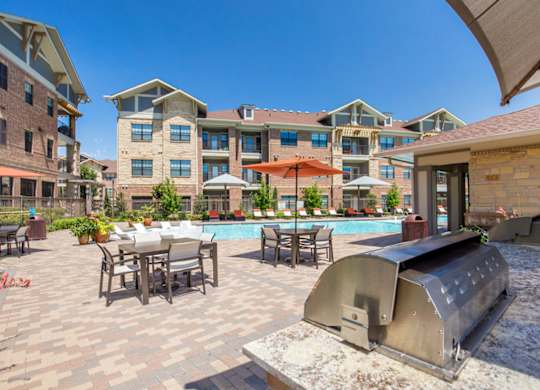 A poolside grilling and dining patio here at Sorrel Fairview with stainless steel grill, stone counter, paver deck, shaded tables with umbrellas, and a bright resort-style pool surrounded by modern brick apartment buildings.