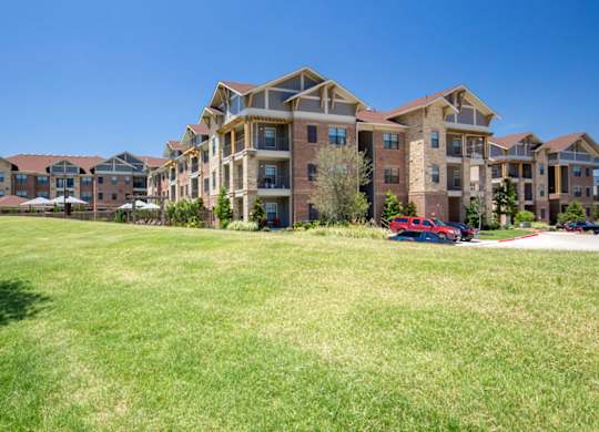 A wide exterior lawn view here at Sorrel Fairview with green open grass leading to multi-story brick and stone apartment buildings with balconies under a clear bright blue sky.