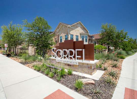A community entrance sign here at Sorrel Fairview with modern monument lettering, landscaped greenery, stone base, and surrounding apartment buildings under a clear blue sky.